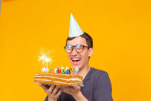 노란색 배경에 그의 손에 축하 수제 케이크를 들고 종이 모자와 안경을 가진 재미있는 긍정적 인 남자의 초상화. 컨셉과 재미와 축하. — 스톡 사진