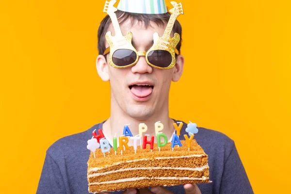 Retrato de un tipo positivo divertido con una gorra de papel y vasos sosteniendo un pastel casero de felicitación en sus manos sobre un fondo amarillo. Concepto y diversión y celebración . — Foto de Stock