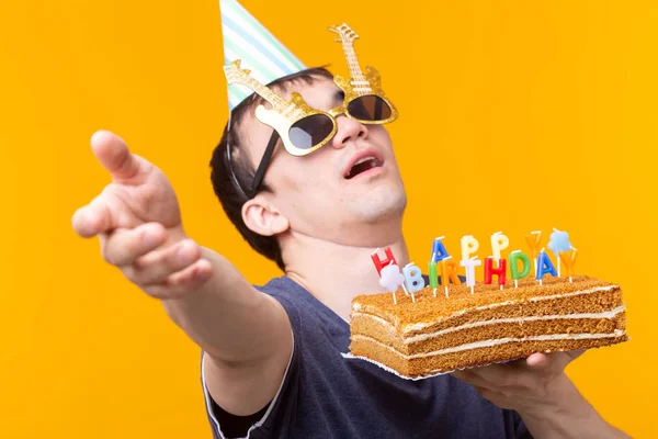 Jovem alegre louco em óculos e chapéus de papel parabéns segurando bolos feliz aniversário de pé em um fundo amarelo. Conceito de parabéns Jubileu . — Fotografia de Stock