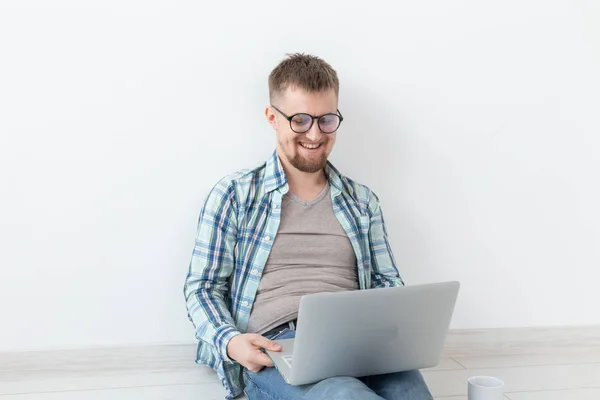 Joven positivo en ropa casual navegando por Internet en busca de una nueva vivienda sentada en el suelo en una habitación vacía. El concepto de encontrar un apartamento a través de Internet y un portátil . — Foto de Stock