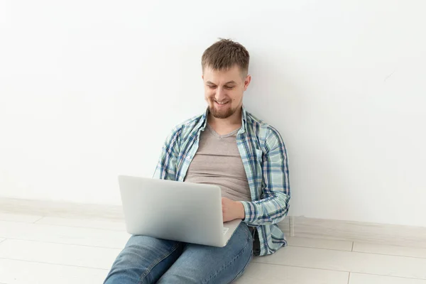 Pozytywny młody człowiek w casual ubrania surfowanie po Internecie w poszukiwaniu nowych mieszkań siedzi na podłodze w pustym pomieszczeniu. Koncepcja znalezienia mieszkania za pomocą Internetu i laptopa — Zdjęcie stockowe