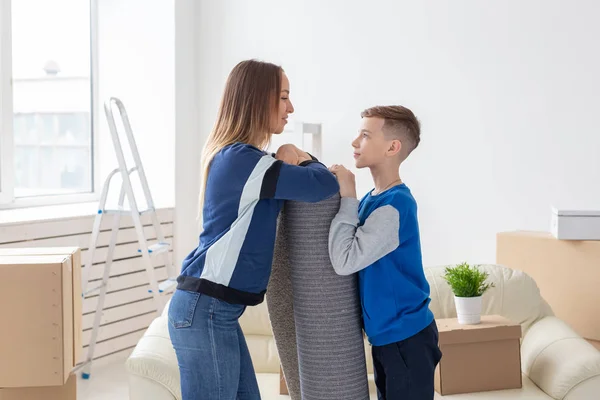 이주, 집안일 및 부동산 개념 - 미소 미혼모와 매력적인 아들이 새 아파트에 퍼뜨리려는 새로운 거실에서 접힌 카펫을 들고 있습니다. — 스톡 사진