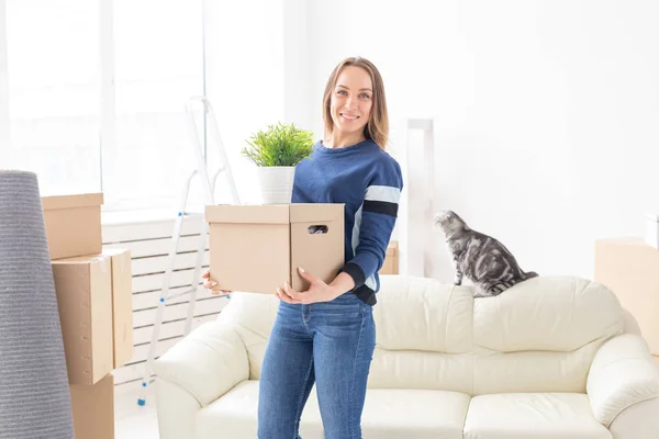 Чарівна молода самотня жінка тримає коробку з речами під час руху, стоячи в новій квартирі поруч зі своїм сірим лопатевим шотландським котом. Концепція переміщення та утеплення . — стокове фото