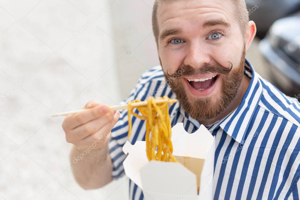 Close-up of an amusing young hipster guy eating chinese noodles with wooden chopsticks sitting in a park outside on a warm summer day. The concept of rest and snacking on the street.