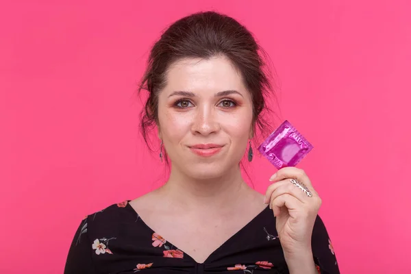 Safe sex, health and contraception concept - a condom in womans hands close up on pink background