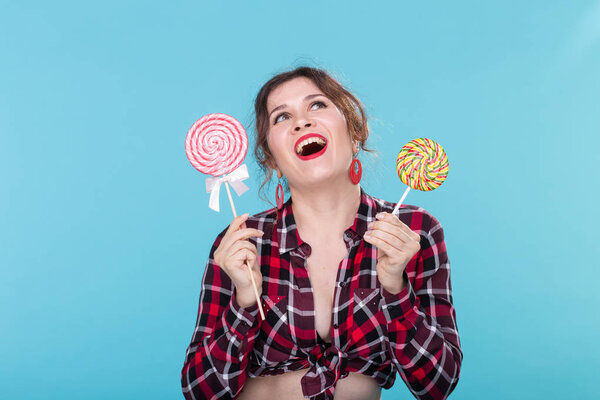 Charming young woman in retro clothes holding colorful lollipops in her hands and licking one posing on blue background. Concept of love for sweets and holidays.