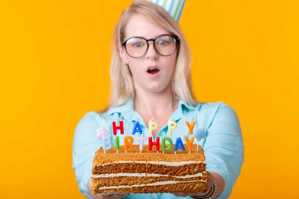 Zabawna pozytywna młoda kobieta trzyma w dłoniach domowy tort z napisem Happy Birthday stwarzających na żółtym tle. Koncepcja świąt i rocznic. — Zdjęcie stockowe