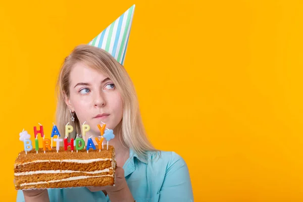 Szalony wesoły młoda kobieta w papier gratulacyjny kapelusz gospodarstwa ciasta szczęśliwy urodziny stojący na żółtym tle z kopią miejsca. Jubileuszowa koncepcja gratulacji. — Zdjęcie stockowe
