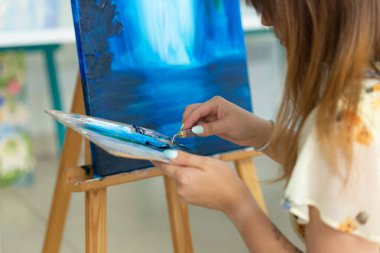 Sanat sınıfı, çizim ve yaratıcılık konsepti - Palet ve boya fırçası ile şövale önünde oturan kız öğrenci, yakın çekim