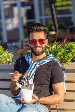 Bıyıklı ve sakallı genç erkek hipster beyaz bir öğle yemeği kutusundan ahşap çubuklarla Çin erişte yiyor. Asya mutfağı konsepti.