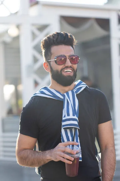 Guapo joven estudiante árabe alegre con un bigote elegante y una barba con jugo en las manos, caminando por la ciudad después de un día de trabajo. Concepto de positivo y descanso . — Foto de Stock