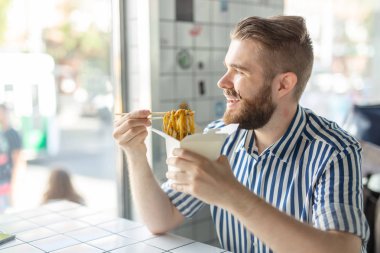 Bir kafede oturan ve pencereye bakarak ahşap çubukları kullanarak Çin erişte yiyen yakışıklı bir genç hipster adam Yan görünümü. Asya mutfağı konsepti.