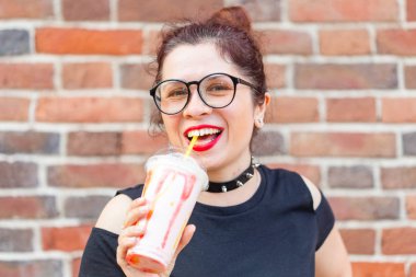 Büyüleyici neşeli genç şık kız bir tuğla duvar arka plan üzerinde bir saman ile milkshake tutan. Bir kafede ve sokakta yaz tatili kavramı.