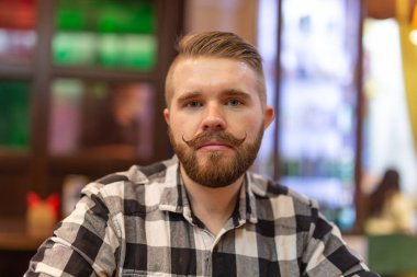 Portrait of a handsome thoughtful young man with a mustache and beard looking at camera in a cafe on a blurred background. The concept of student or stylish men. clipart