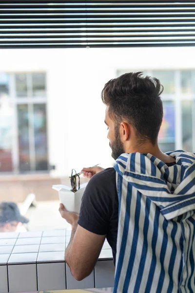 Yakışıklı genç hipster adam bir kafede oturan ve pencereye bakarak ahşap çubukları kullanarak Çin erişte yeme. Asya mutfağı konsepti.