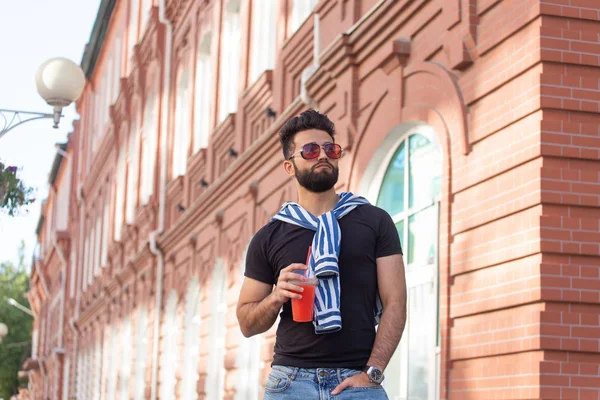 Retrato de un joven árabe alegre positivo con un vaso de jugo con una pajita mientras camina por la ciudad en un cálido día soleado de verano. El concepto de descanso después del estudio y el trabajo los fines de semana . — Foto de Stock