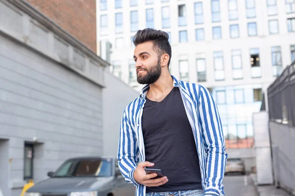 Joven y elegante árabe hipster hombre con barba charlando en las redes sociales utilizando un teléfono inteligente e internet inalámbrico en un cálido día de verano . — Foto de Stock