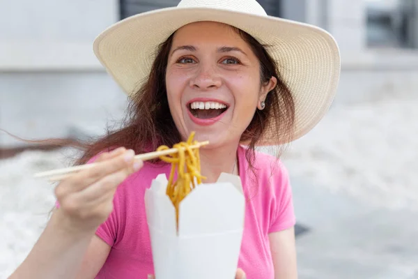 Mladá pozitivní dívka v klobouku sní čínské nudle s dřevěnými hůlky na oběd s sebou. Koncept vysoce kalorických zdravých potravin. — Stock fotografie