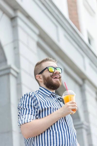 Retrato de un joven hipster borroso y elegante con barba y bigote sosteniendo jugo de naranja fresco en sus manos. Concepto de snack saludable . — Foto de Stock