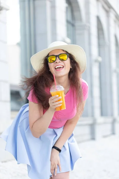 Portret wesołej pozytywnej młodej kobiety w kapeluszu i szklankach z sokiem w dłoniach spacerowych po mieście w słoneczny ciepły letni dzień. Pojęcie beztrofree nastroju. — Zdjęcie stockowe