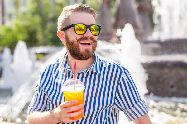 Schattig stijlvolle jonge mannelijke hipster met een snor en een baard en het dragen van glazen drinken sap met een rietje terwijl zittend in een stadspark tegen de achtergrond van een fontein. Zomer vakantie concept. — Stockfoto