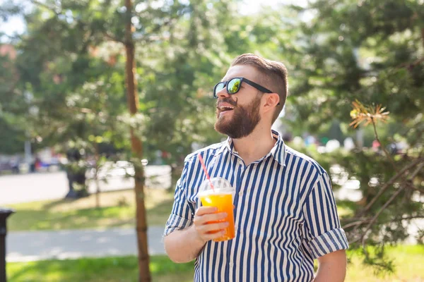 Retrato de un joven positivo y alegre con un vaso de jugo con una pajita mientras camina por el parque en un cálido día soleado de verano. El concepto de descanso después del estudio y el trabajo los fines de semana . — Foto de Stock