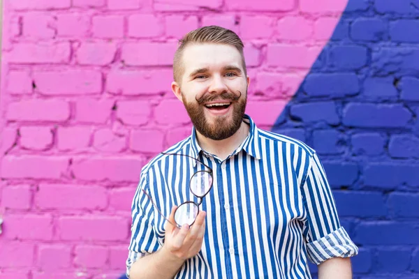 Retrato de joven alegre positivo elegante con bigote y barba y gafas en las manos posando sobre fondo de pared de ladrillo azul-violeta . — Foto de Stock