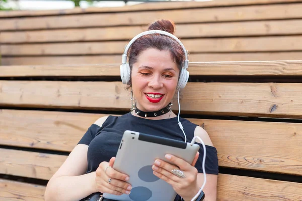 Beautiful young female student is studying using a tablet and headphones while sitting in a park on a wooden bench. Recreation and study concept. — Stock Photo, Image