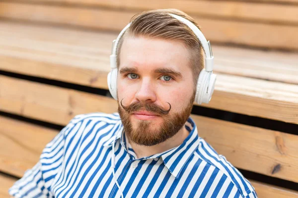 Close-up portret van een serieuze jonge studente die luistert naar zijn favoriete online radio met hoofdtelefoon terwijl je in een park zit op een zonnige warme zomerdag. — Stockfoto