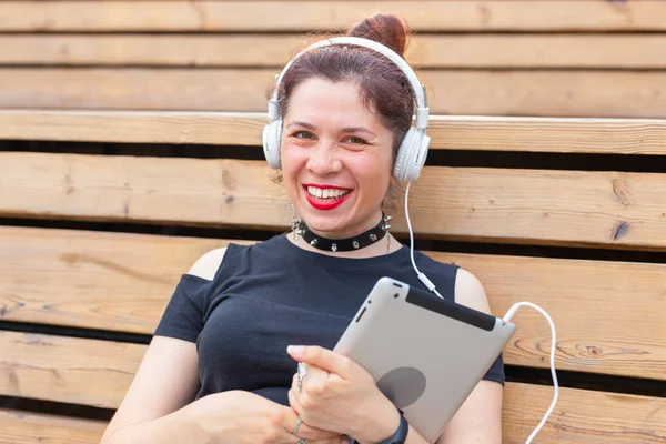 Beautiful young female student is studying using a tablet and headphones while sitting in a park on a wooden bench. Recreation and study concept. — Stock Photo, Image