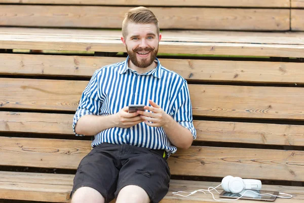 Feliz joven estudiante hipster masculino con bigote y barba escribiendo sms mensaje a sus amigos descansando después de estudiar en el parque sentado en un banco de madera con una tableta y auriculares — Foto de Stock
