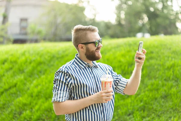 Vista lateral de un joven elegante con gafas y una barba con un batido en las manos hablando en video con sus amigos usando un teléfono inteligente e Internet gratuito de alta velocidad en el parque en un cálido día de verano — Foto de Stock