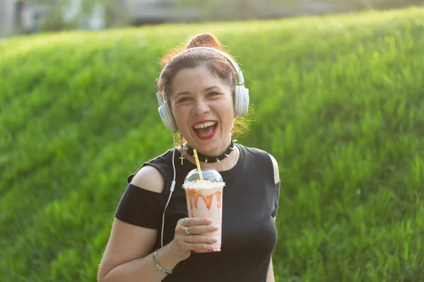 아름다운 관능적 인 젊은 여성은 헤드폰으로 음악을 듣고 화창한 저녁에 공원을 산책하는 동안 그녀의 손에 밀크 셰이크를 들고있습니다. 주말 휴양지 컨셉. — 스톡 사진