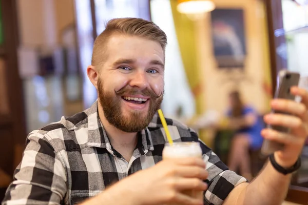 Un joven elegante con bigote y barba está viendo una red social usando un teléfono inteligente mientras está sentado en un café un fin de semana. El concepto de dependencia de las redes sociales . — Foto de Stock