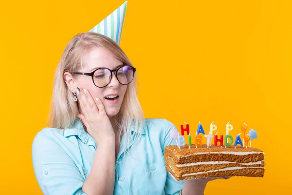 Divertente giovane donna positiva tiene in mano una torta fatta in casa con l'iscrizione buon compleanno in posa su uno sfondo giallo. Concetto di vacanze e anniversari . — Foto Stock