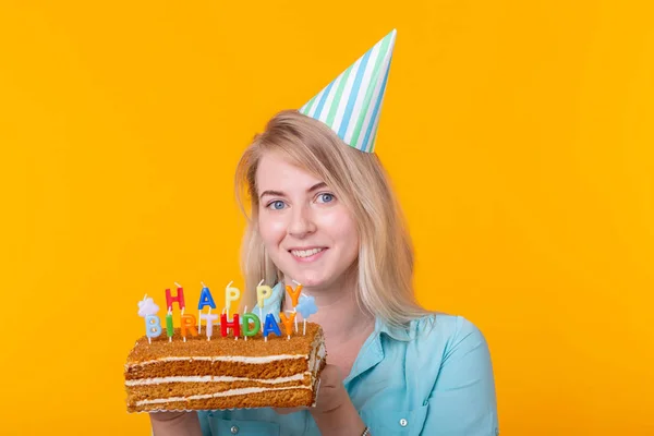 Позитивна смішна молода жінка з шапочкою і домашнім тортом в руках позує на жовтому тлі. Концепція ювілею та дня народження . — стокове фото