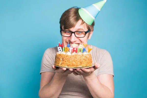 Drôle jeune homme fou dans un chapeau de papier de salutation veut mordre un morceau de gâteau de félicitations. Concept de plaisanterie et salutations d'anniversaire. Espace de copie — Photo