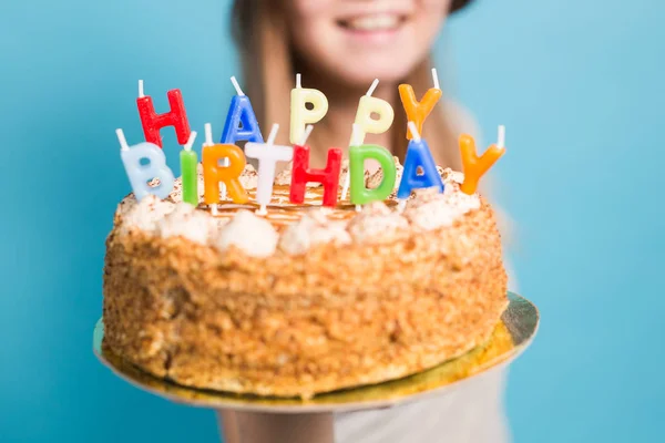 De cerca divertida chica positiva en gafas y sombrero de papel de saludo sosteniendo un pastel de cumpleaños feliz en sus manos de pie sobre un fondo azul . —  Fotos de Stock