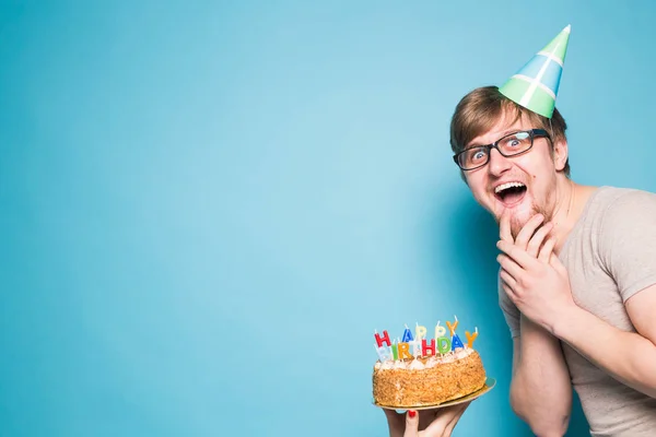 Crazy vicces fiatalember a szemüveg és a papír gratuláló kalap gazdaság sütemények boldog születésnapot állva a kék háttér copyspace — Stock Fotó