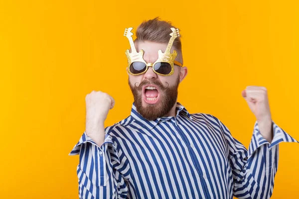 Joven positivo con gafas en forma de guitarras se regocija sobre un fondo amarillo. El concepto de celebración y fiestas . — Foto de Stock