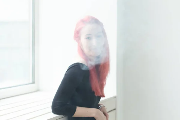 Youth and addiction concept - young red haired woman smoking vape near the window