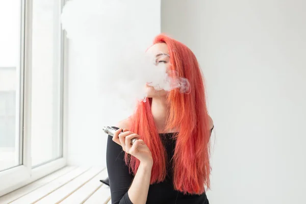 Youth and addiction concept - young red haired woman smoking vape near the window
