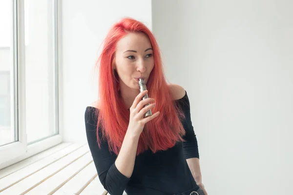 Youth and addiction concept - young red haired woman smoking vape near the window — Stock Photo, Image