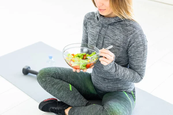 Sport, healthy lifestyle and people concept - young woman with salad and a dumbbell sitting on the floor