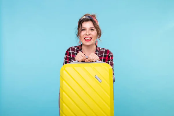 Joven mujer sonriente positiva con una camisa a cuadros que sostiene una maleta amarilla posando sobre un fondo azul. Concepto de turismo y viajes — Foto de Stock