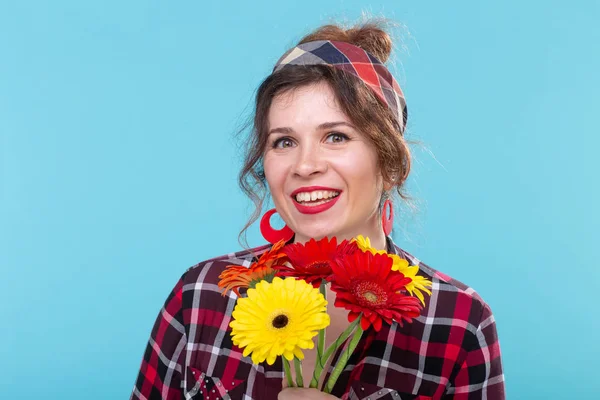 격자 무늬 셔츠와 파란색 배경 위에 포즈를 취하는 아름다운 밝은 거베라 꽃을 스니핑 붕대에서 아름다운 긍정적 인 젊은 여성. 선물과 인사의 개념. — 스톡 사진