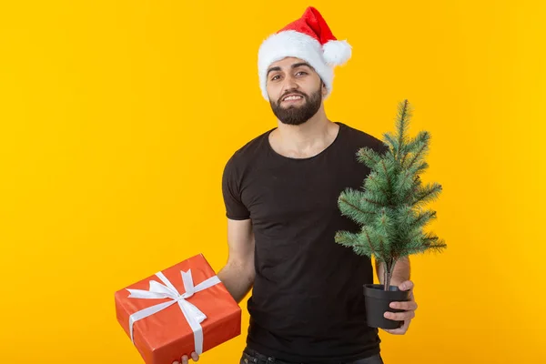 Knappe jonge man met een rode geschenk doos en kerstboom in zijn handen poseren in een nieuwe jaar GLB op een gele achtergrond. Merry Christmas en Happy New Year groeten concept. — Stockfoto