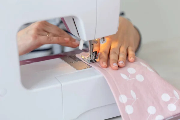 Cerrar las manos de una joven costurera coser un nuevo producto en una máquina de coser sosteniendo un paño rosa. El concepto de crear productos de costura a pedido . — Foto de Stock