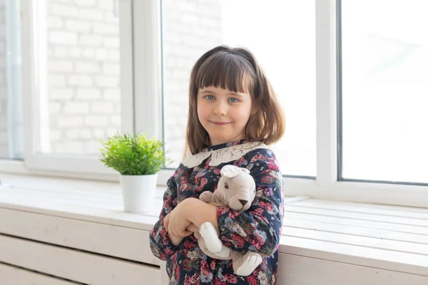 Porträtt av en söt snäll liten flicka i en blomma klänning med en favorit mjuk kanin leker leksak i händerna stående nära fönstret. Konceptet med Childs rum och relaterade produkter. — Stockfoto