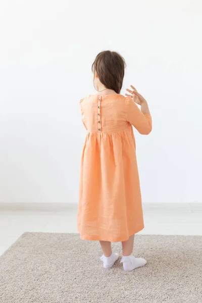 Vacker liten flicka i en persika klänning poserar mot en vit vägg. Begreppet barndesigner kläder. — Stockfoto